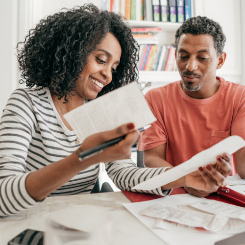 A smiling woman and man sit at a table reviewing receipts and documents. The woman holds a pen and notebook while the man gestures with a paper, suggesting they are planning their family budget.