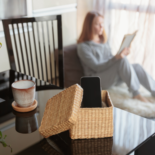 A cozy home scene with a smartphone tucked away in a basket on a table while a women relaxes, reading a book in the background, capturing a peaceful digital detox moment.