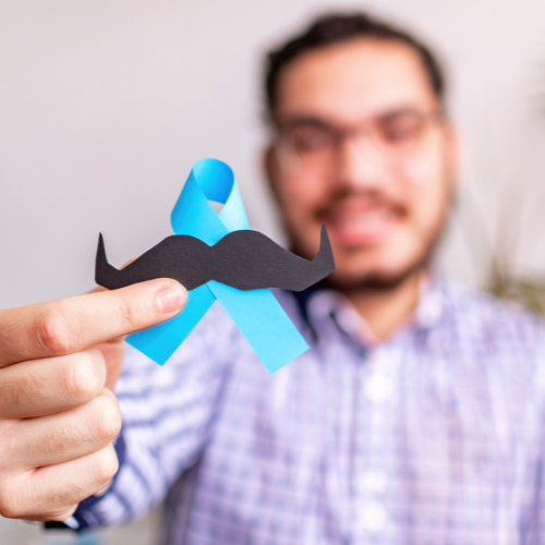 Personne tenant une moustache en papier et un ruban bleu de sensibilisation, symbolisant le soutien à Movember et à la sensibilisation à la santé des hommes.