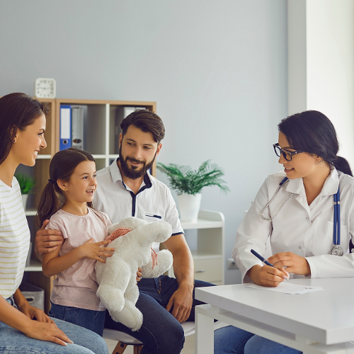 A young girl holding a stuffed animal sits with her parents during a check-up with a female doctor in a bright medical office.
