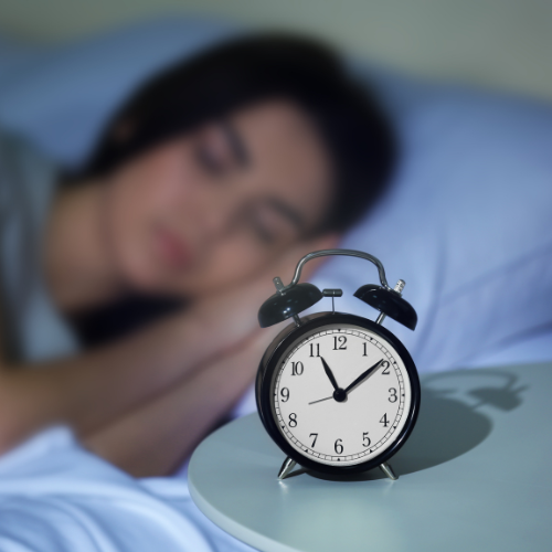 A black alarm clock showing 11:55 sits on a bedside table, with a woman sleeping peacefully in the blurred background, evoking calm and rest.