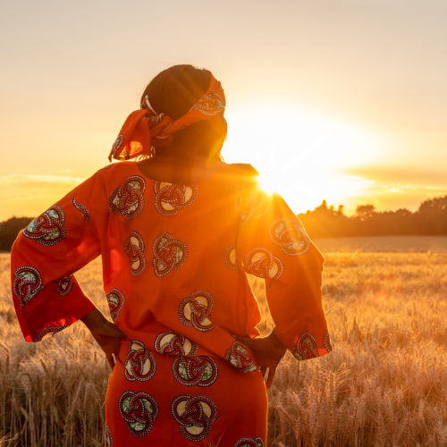 Person dressed in an orange shirt with traditional patterns standing in a wheat field at sunset, symbolizing Orange Shirt Day and honouring Indigenous children and survivors.